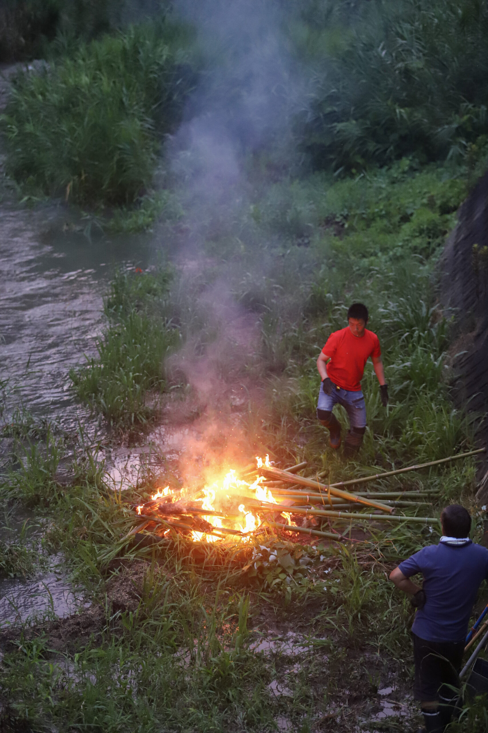 河畔の草地で焚き火を囲む2人の男性。夕暮れ時に炎が明るく輝き、周囲の緑豊かな自然環境とのコントラストが印象的なキャンプシーンです。